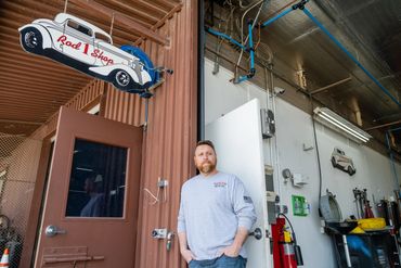 A man with a beard stands in the doorway of a shop, beside a sign shaped like a classic car that rea