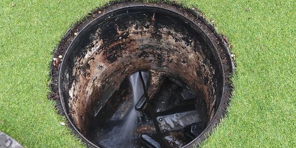 Open manhole with water flowing inside, surrounded by green grass.