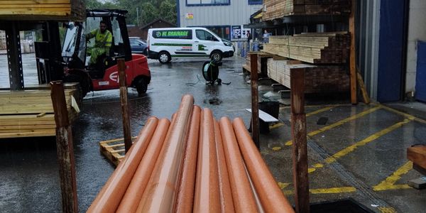 Stack of orange pipes in a wet lumberyard with workers and vehicles.