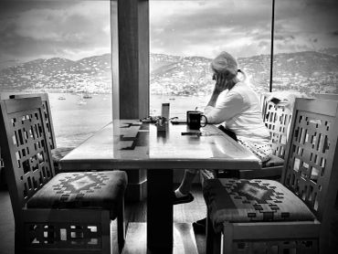 A woman sits alone at a wooden table overlooking a scenic waterfront through large windows.