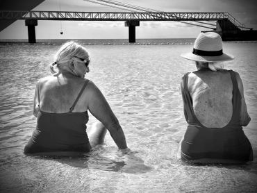 Two elderly women relaxing in shallow water by a bridge, one wearing a hat.