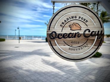 Wooden sign for Ocean Cay Trading Post with beach and ocean backdrop.