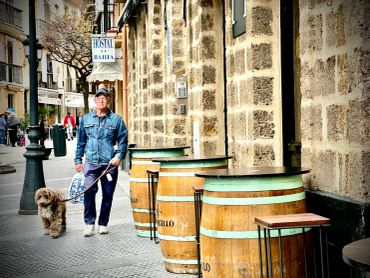 Man walking a dog past wooden barrels outside a stone building.