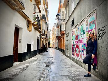 A woman stands casually on a narrow, urban street with colorful graffiti art on the wall.