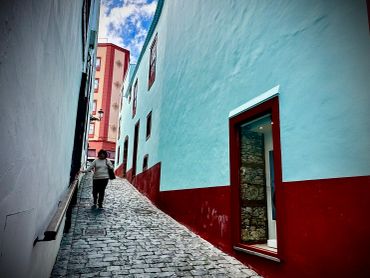 Person walking up a narrow cobblestone street between white and red buildings.