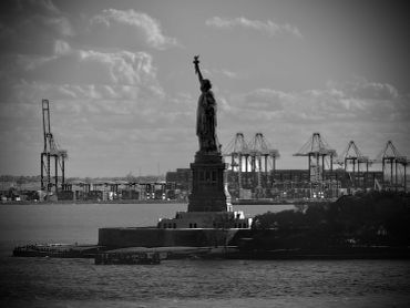 Black and white photo of the Statue of Liberty with industrial cranes in the background.