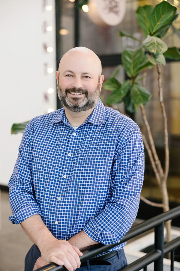 Smiling man in a blue checkered shirt leaning on a railing indoors.