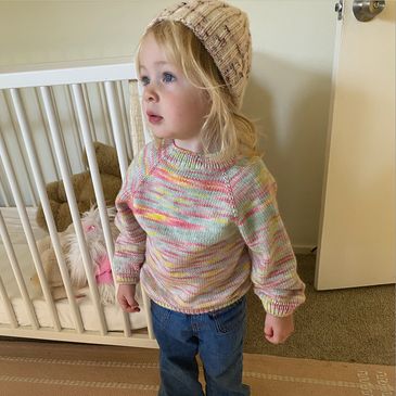 A toddler wearing a knit hat and colorful sweater stands indoors near a crib.