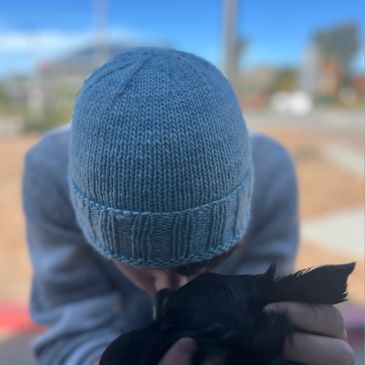 Person in a blue knit hat holding and kissing a small black dog.