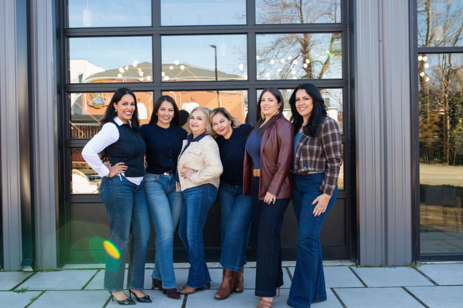 Six women stand smiling in front of a large windowed door, dressed in casual and business casual attire.
