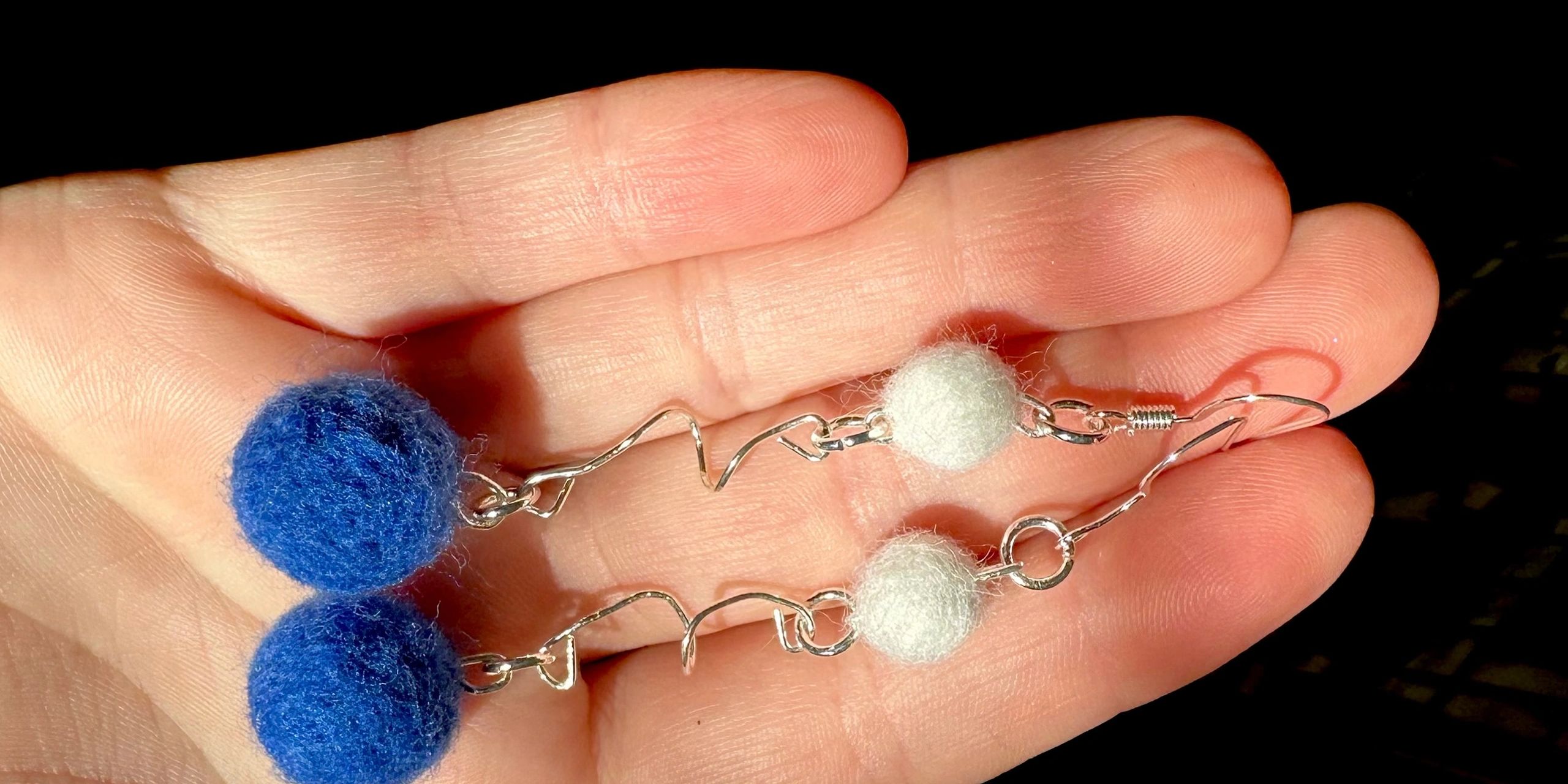 Hand holding handmade earrings with blue and white felt balls and silver hooks.