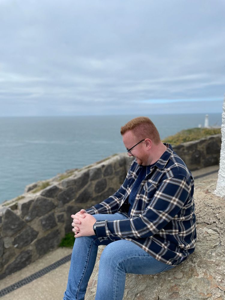 Man in glasses sitting on a rock near the ocean, smiling and looking down.