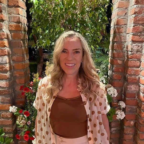 Smiling woman standing between brick arches with greenery and flowers behind her.