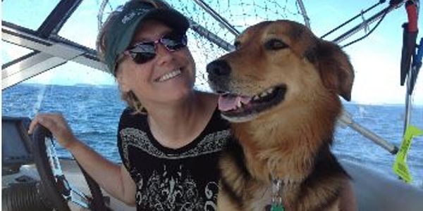 Woman wearing sunglasses smiles while steering a boat with a happy dog beside her.