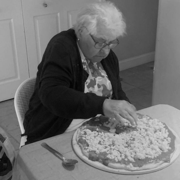 Elderly woman spreading cheese on a large pizza dough.