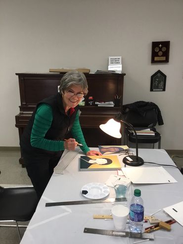Elderly woman painting a portrait in a well-lit room.