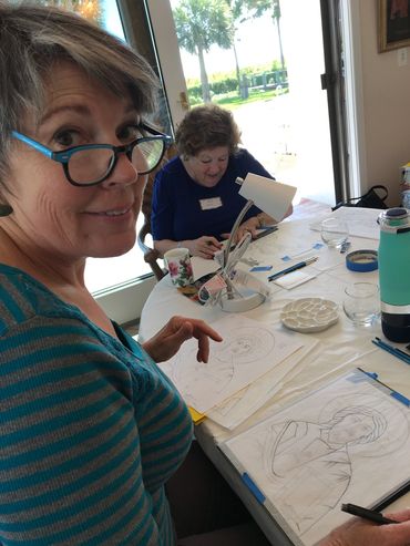 Two women drawing sketches at a sunlit table indoors.
