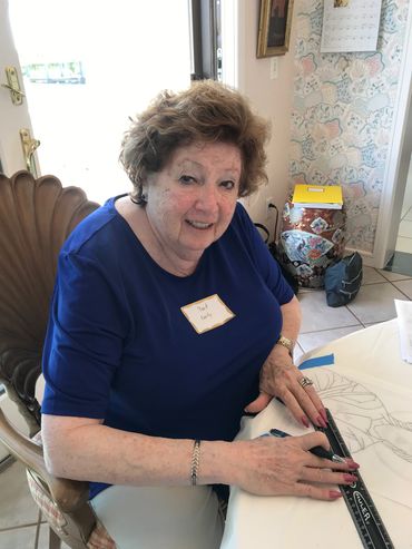 Elderly woman in blue shirt sitting at a table, smiling, with a ruler and drawing materials.