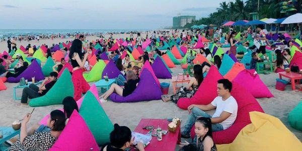 Crowded beach with people relaxing on colorful bean bags near the shore.