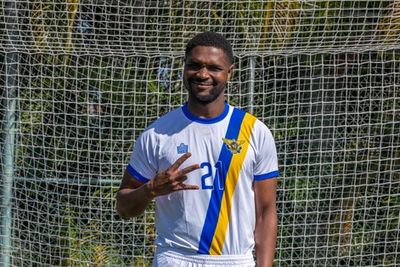 Soccer player in white and blue uniform posing with a peace sign on the field.