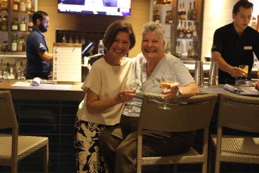 Two women enjoying drinks and smiling at a bar with bartenders in the background.