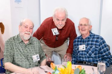 Three elderly men smiling at a table with name tags and flowers.