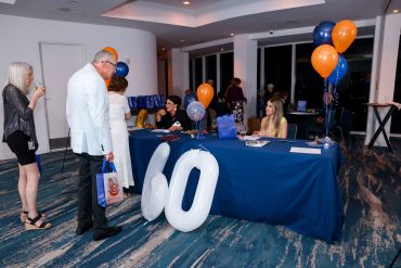 A man in a white blazer examines large white balloon numbers '60' at an event registration table.