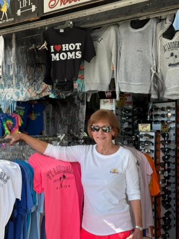 Woman smiling inside a souvenir shop with colorful t-shirts and sunglasses.