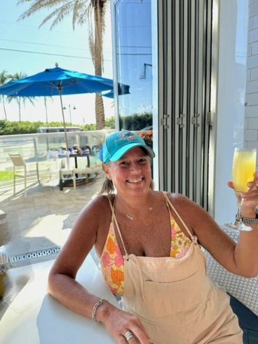 Woman smiling with a drink in hand at an outdoor seating area.