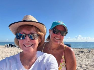 Two women smiling on a sunny beach with clear blue sky and ocean behind them.
