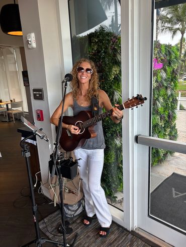 Woman playing guitar and singing indoors near a glass door.