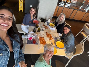 Five women smiling around a table with pizzas at a casual restaurant.