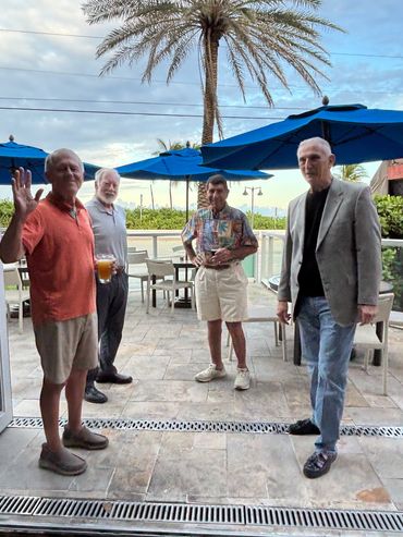Four elderly men stand on a patio with palm trees and blue umbrellas in the background.