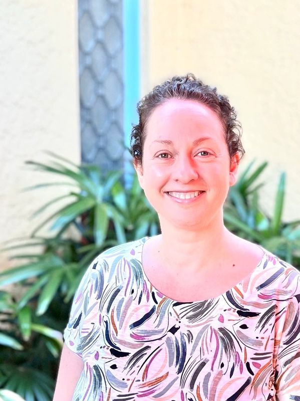 Smiling woman in a colorful patterned shirt outdoors with greenery in the background.