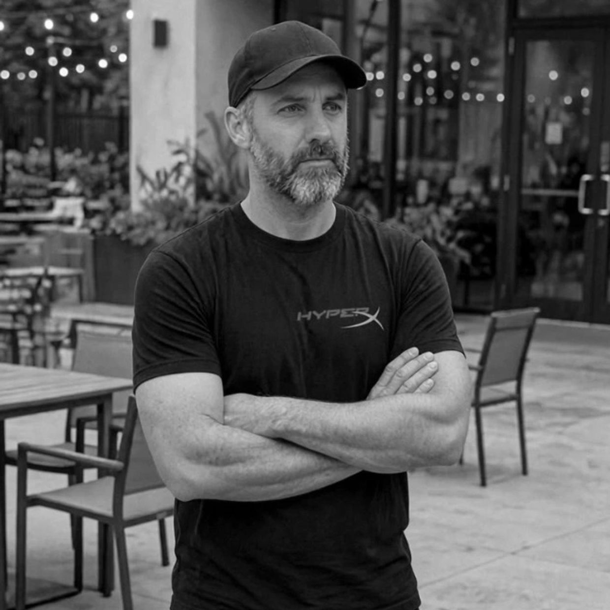 Man with beard and cap standing outdoors with arms crossed.