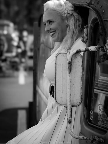 Woman with blonde hair leaning back against rustic car in black and white.
