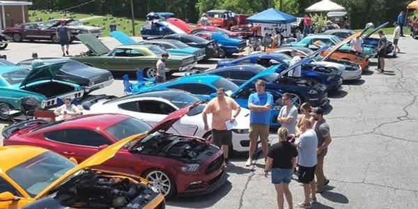 Group of people admiring cars with hoods open at an outdoor car show.