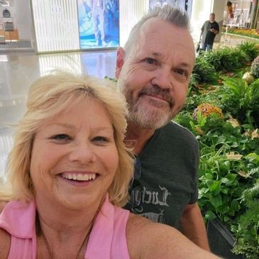 Smiling couple taking a selfie in a mall with lush greenery behind them.