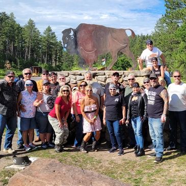 Group of bikers posing in front of a large metal buffalo statue outdoors.