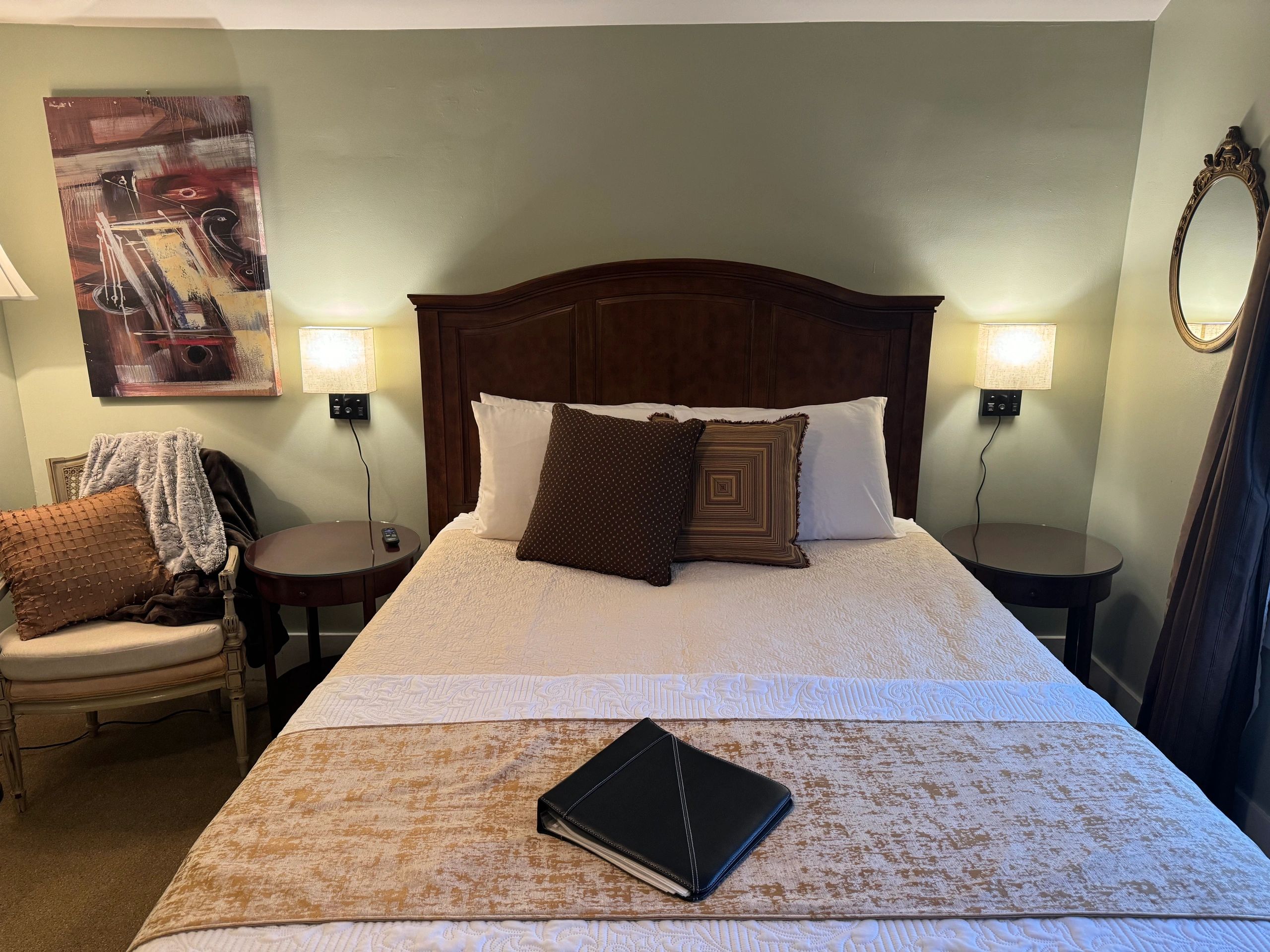 A neatly made bed with a dark wood headboard, white and brown bedding, and two nightstands with lamps, in a room with green walls and a painting.