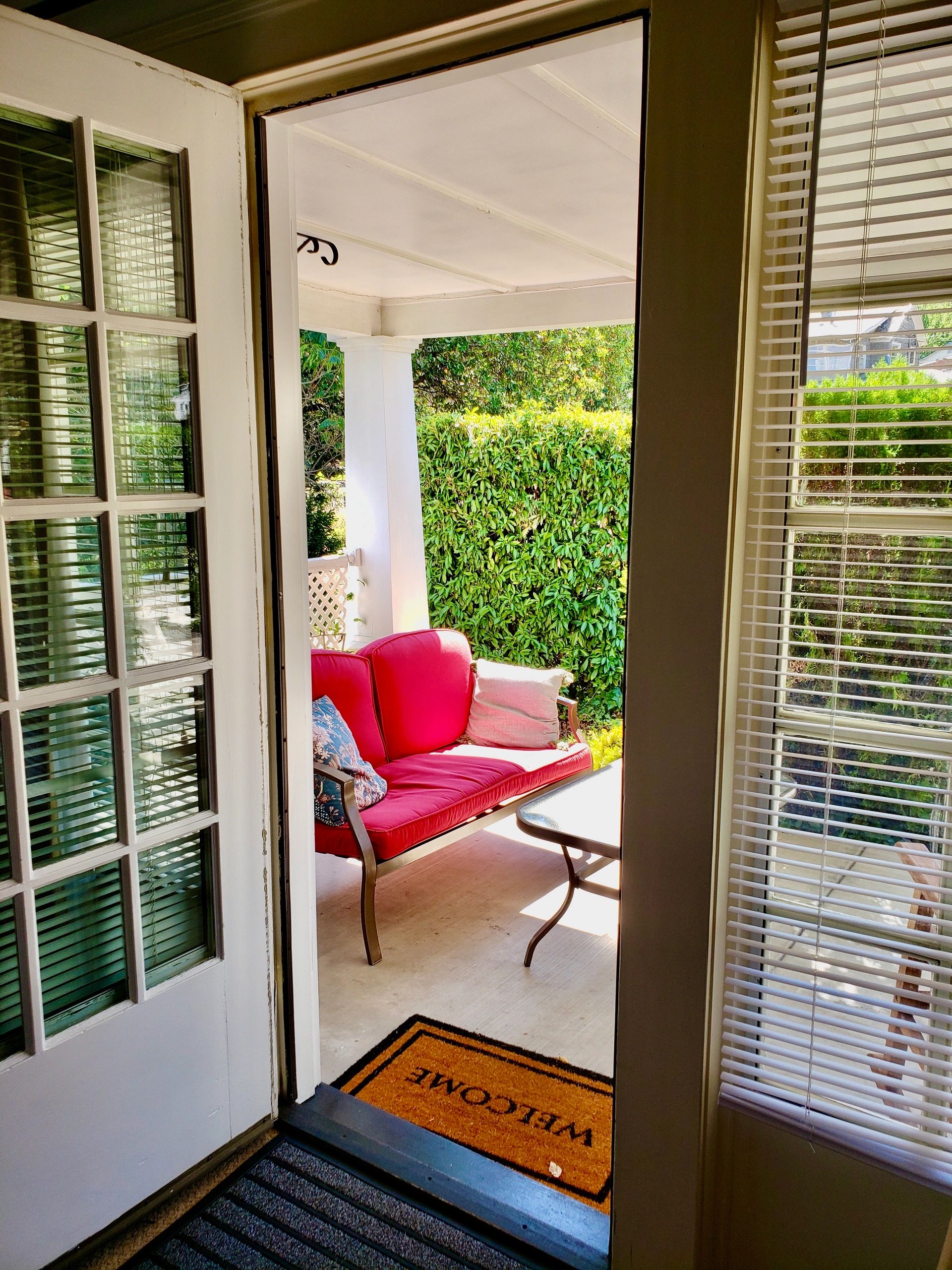A doorway leading to a small porch with a red couch and a hedge in the background.