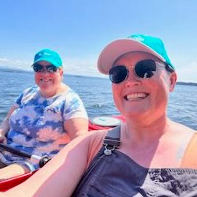 Two women sitting in kayaks on a lake. They are wearing sunglasses and hats and are smiling. 
