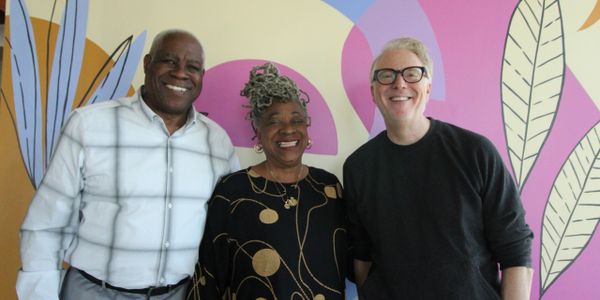Three smiling adults standing in front of a colorful wall mural.