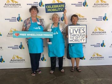 Three women in blue aprons at Free Wheelchair Mission event holding signs.