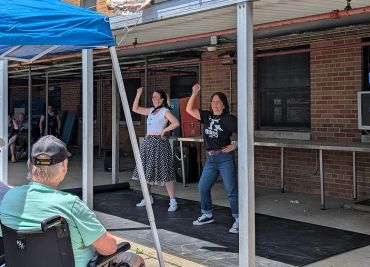 Two women dancing outdoors while a seated man watches.