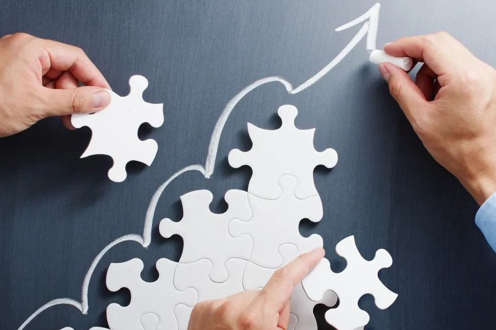 Hands assembling puzzle pieces with an upward arrow drawn on a chalkboard.