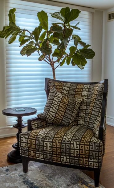 Elegant patterned armchair with cushion beside a small wooden table and a tall leafy plant.