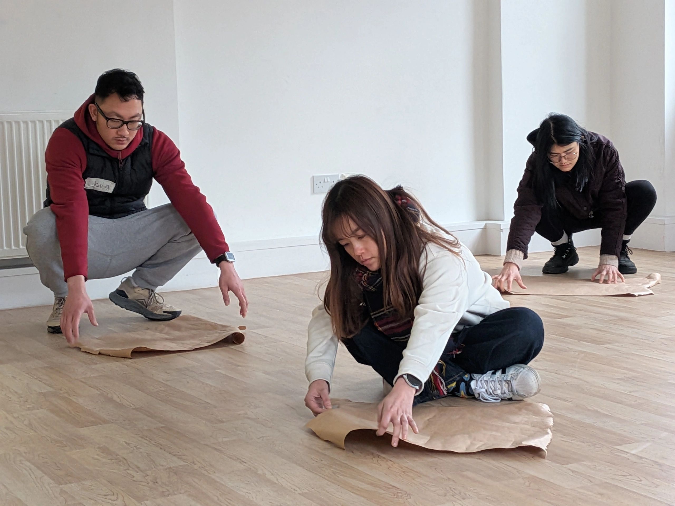 Three people crouched on the floor, exploring breath in a brown paper puppet. 