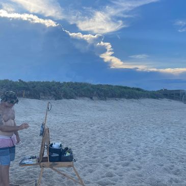 A man painting on a beach under a cloudy sky.