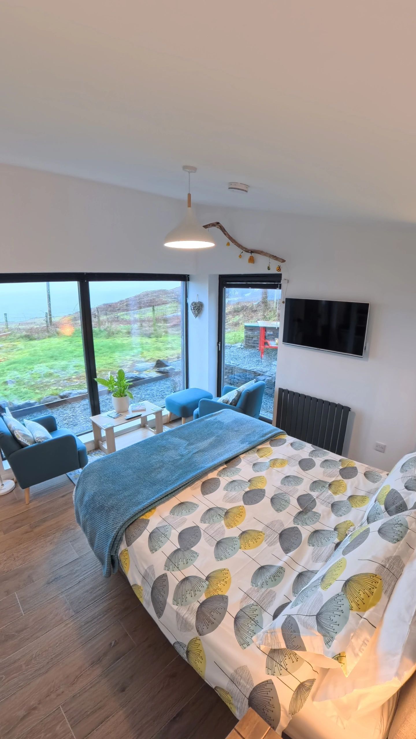 Cozy bedroom with large windows overlooking a green outdoor view, featuring blue chairs and a patterned bedspread.